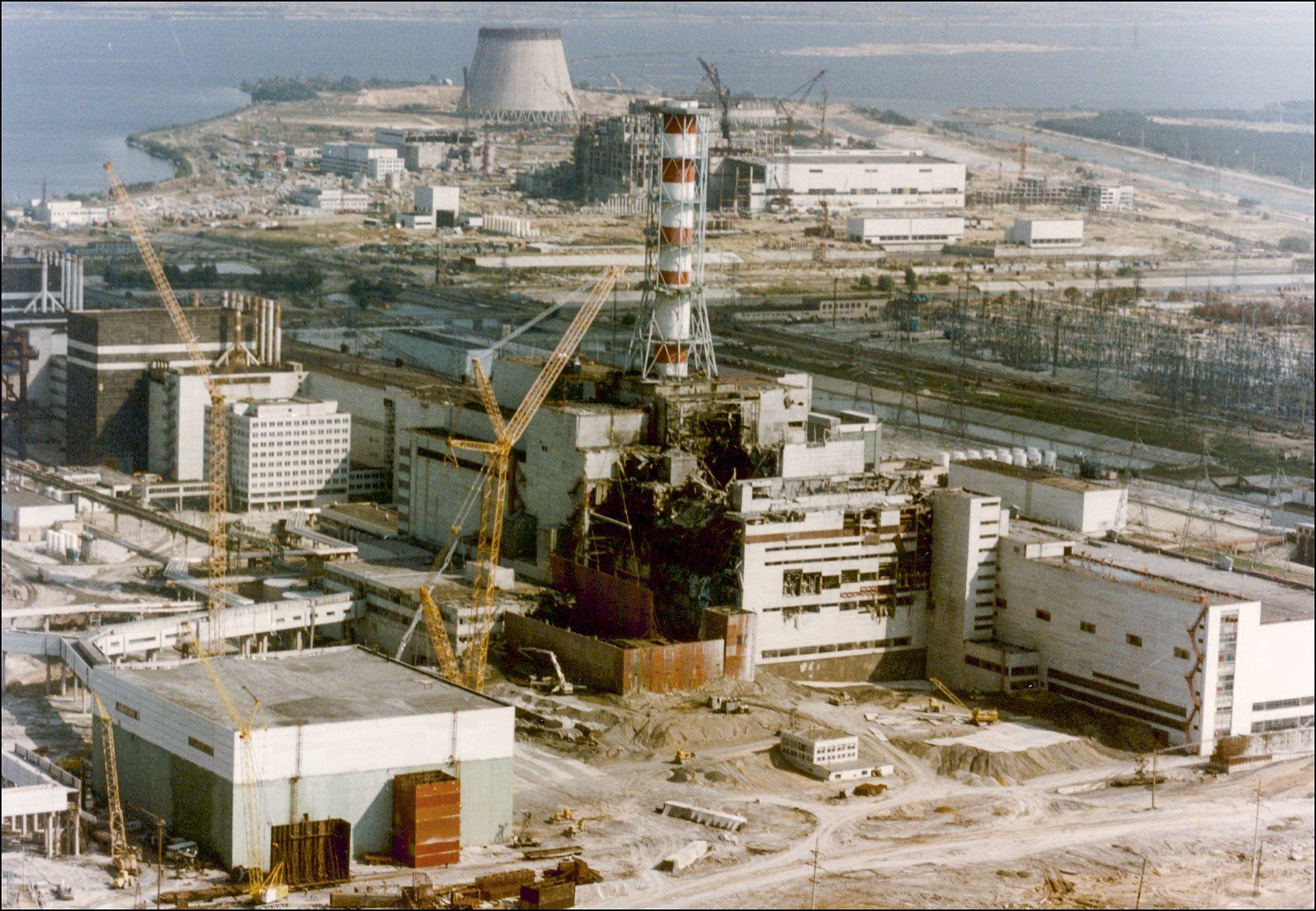 Vista da Usina Nuclear de Chernobyl em 29 de abril de 1986, tr&ecirc;s dias ap&oacute;s a explos&atilde;o. O pr&eacute;dio que abriga o reator quatro est&aacute; gravemente danificado, com destro&ccedil;os enegrecidos abaixo de uma chamin&eacute; vermelha e branca