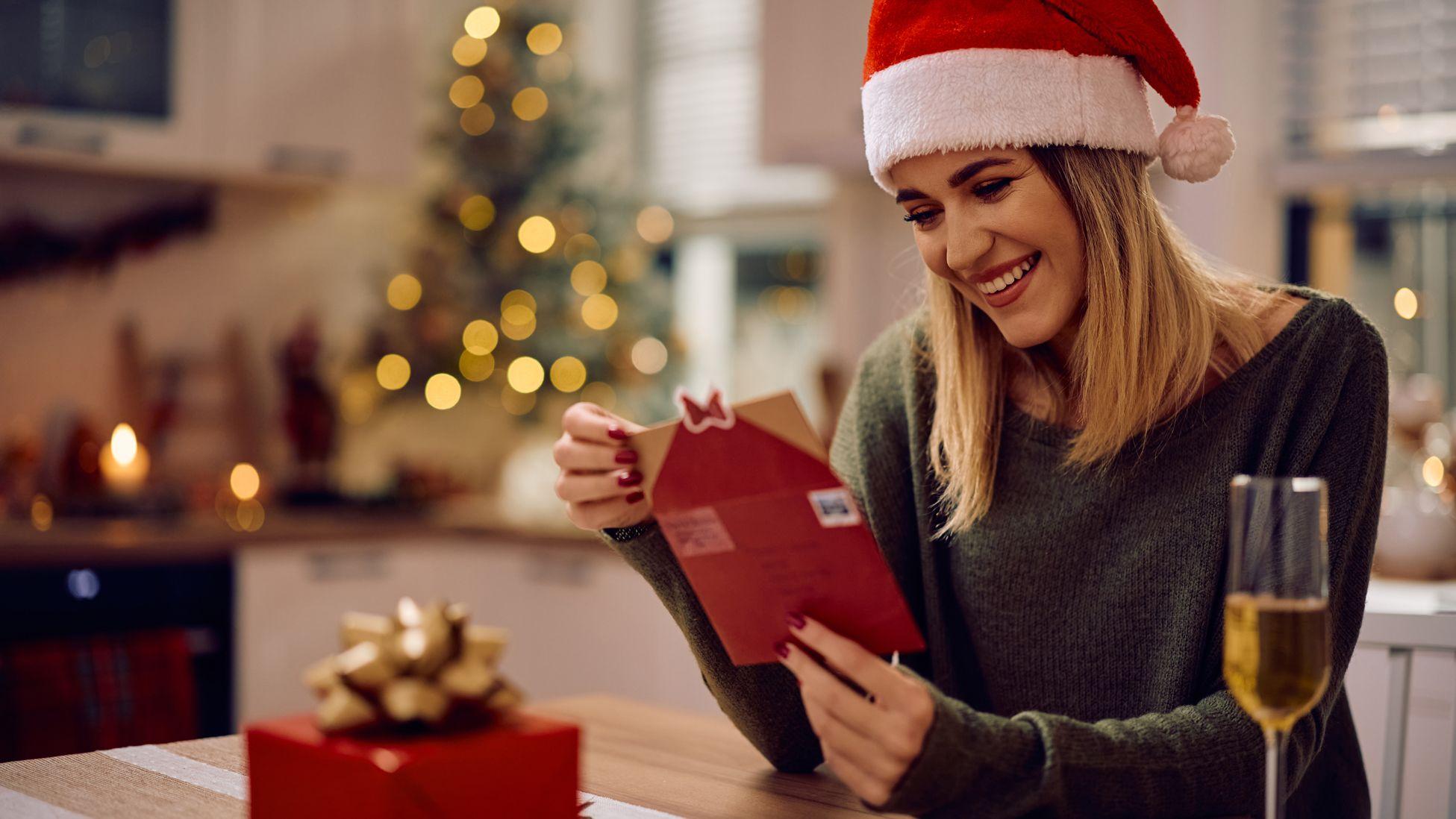 Una mujer con un gorro de Papá Noel abriendo un sobre con una tarjeta de Navidad y al fondo un árbol navideño.