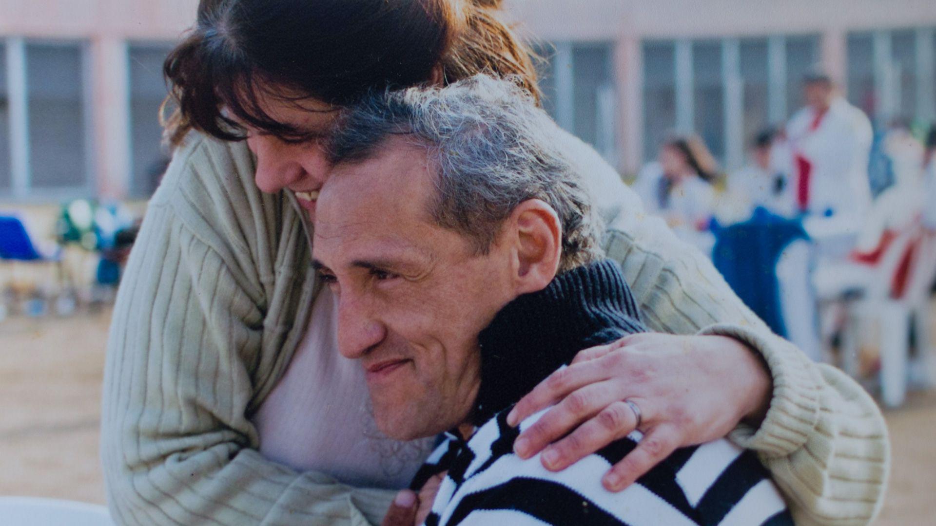 Alejo está sentado y luce complacido mientras Andrea, que está de pie, lo abraza por el costado, le pone la mano izquierda en el hombro y sonríe mientras junta su cabeza con la de él.