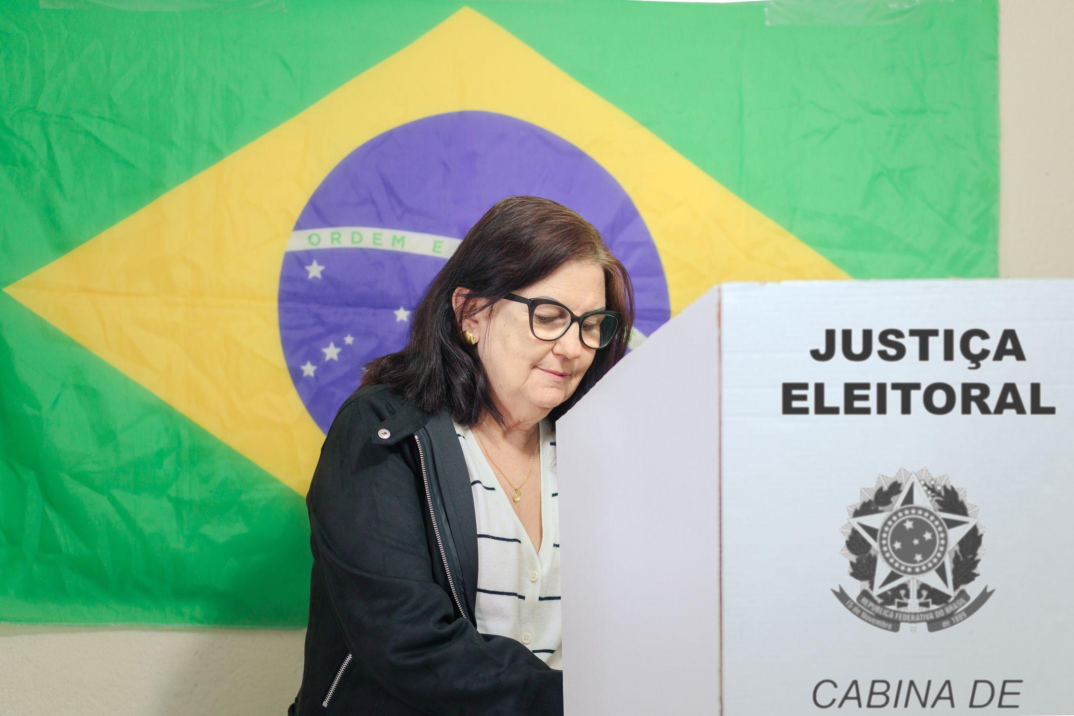 Mulher votando em se&ccedil;&atilde;o eleitoral, com a bandeira do Brasil ao fundo 