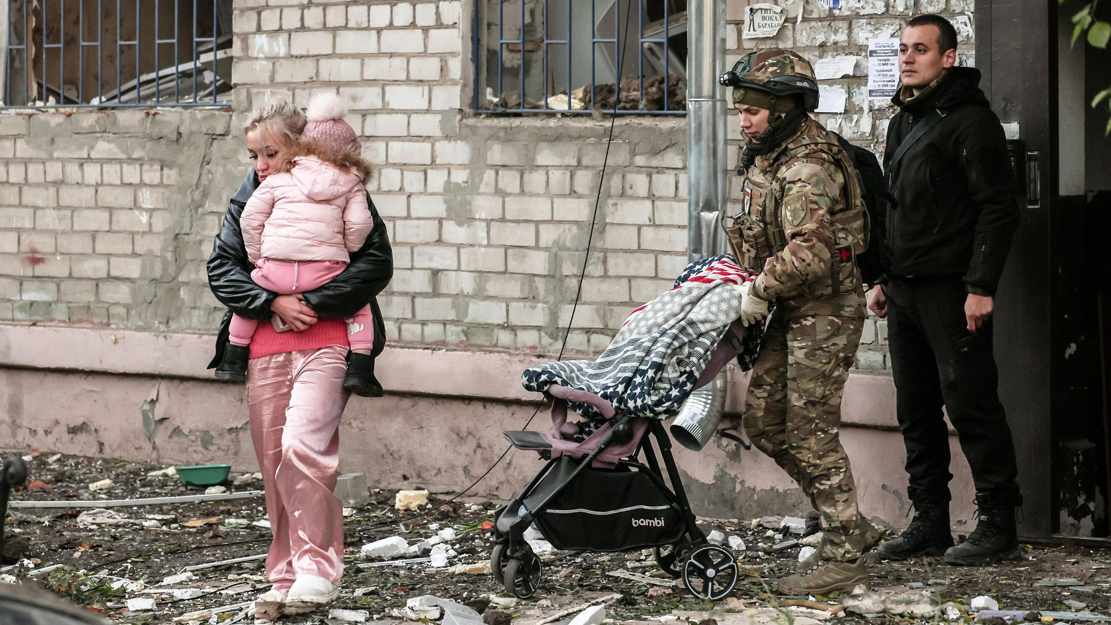 Una mujer, con un niño vestido con pantalones, chaqueta y gorro de lana rosas, sale de un edificio con escombros en el suelo. Un soldado uniformado empuja un cochecito y un hombre permanece de pie junto a la puerta del edificio.