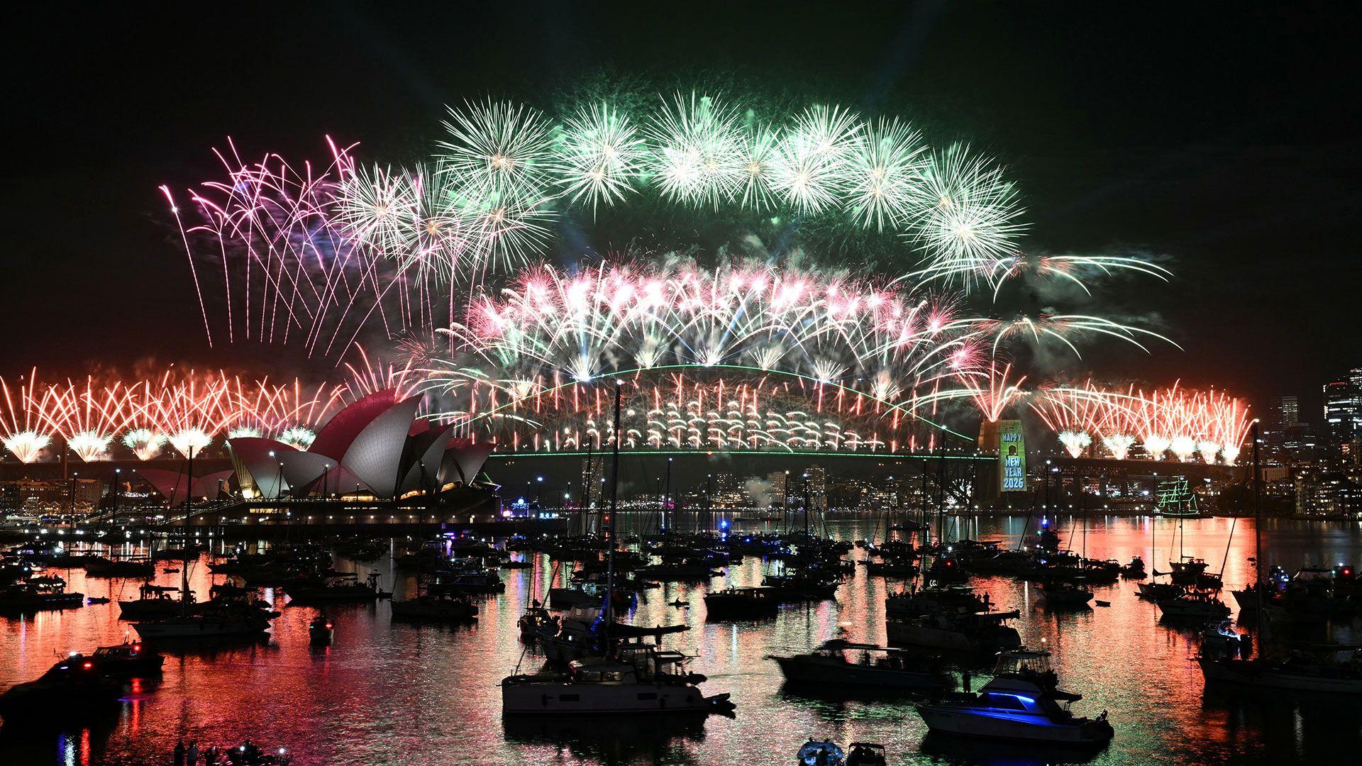Los fuegos artificiales iluminan el cielo nocturno sobre el Puente de la Bahía de Sídney y la Ópera de Sídney durante las celebraciones de Año Nuevo en Australia. Numerosas embarcaciones se encuentran amarradas en el puerto, frente al espectáculo pirotécnico.
