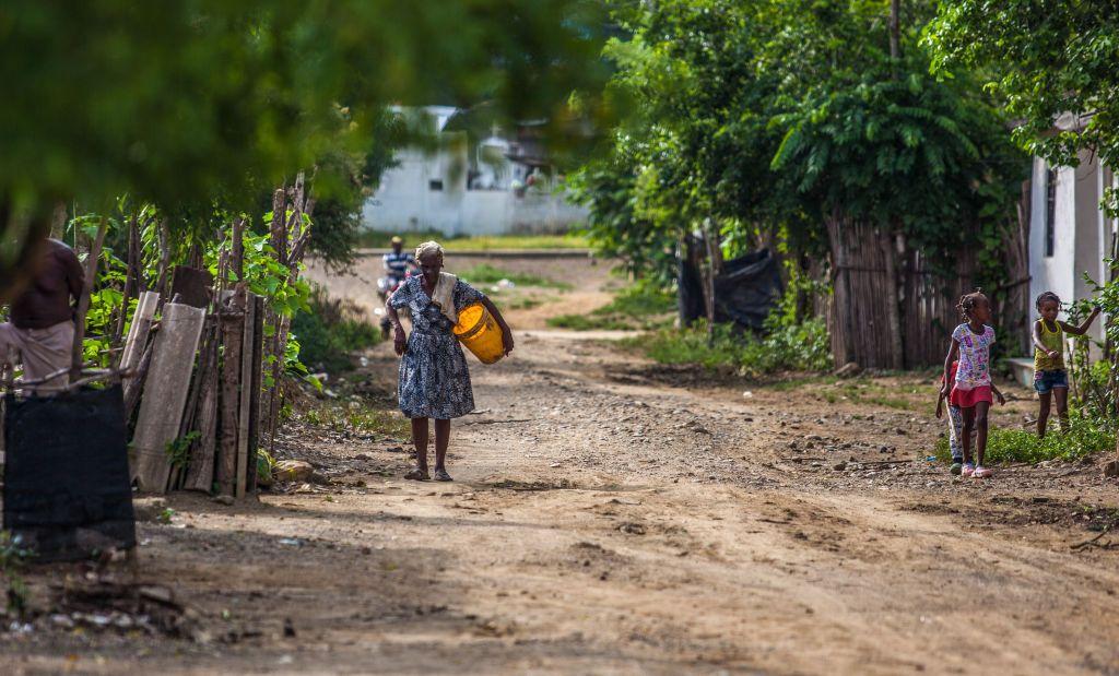 Una mujer afrodescendiente camina por una calle de tierra cargando un balde amarillo. Hay otros niños en la calle y un hombre en bicicleta en el fondo. Hay frondosa vegetación a lado y lado.