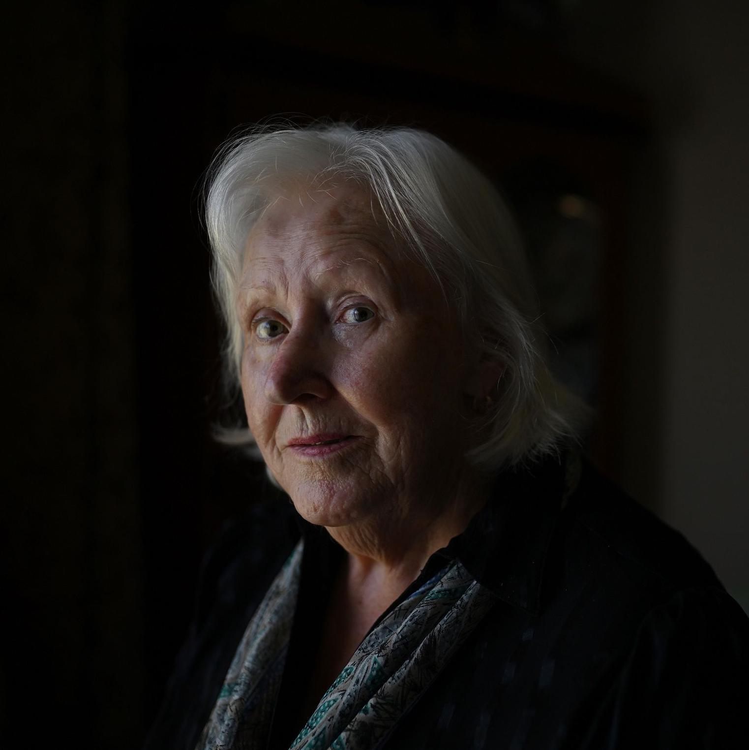 Starija žena sa srednje dugačkom sedom kosom stoji u dnevnoj sobi. Vidimo joj glavu i ramena. Nosi tamnu jaknu i šareni svileni šal. U pozadini, koja je van fokusa, vidimo sliku koja visi na zidu dnevne sobe i ormar.