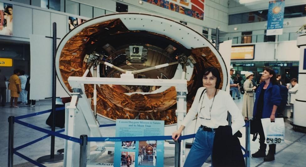 Michele Dougherty, de 30 años, con un top blanco y vaqueros azules, gafas colgando del cuello y una chaqueta negra colgada del brazo. Está de pie delante de una parte redonda de una nave espacial, en parte blanca pero con partes de lámina de cobre. Está delimitada por una barandilla azul. La escena parece transcurrir en un museo, o tal vez en un aeropuerto, y hay gente alrededor.