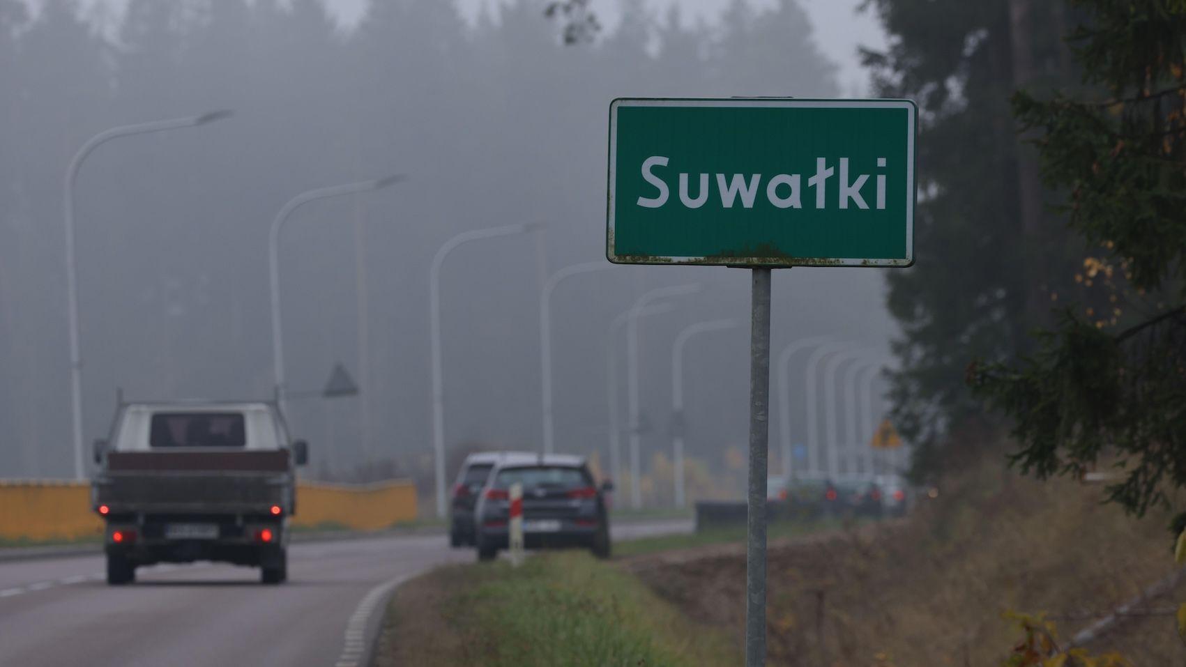 兩輛汽車駛過一座小鎮，道路上有綠色的蘇瓦烏基（Suwałki）限制標誌。背景是一些樹木和路燈。