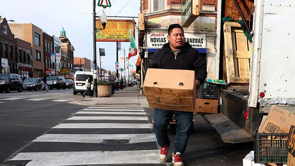 Un vendedor ambulante descarga varias cajas de frutas y vegetales de un camión estacionado en una esquina de la calle 26 en Little Village, Chicago. En el fondo se ve una bandera de México y varios avisos de almacenes en español