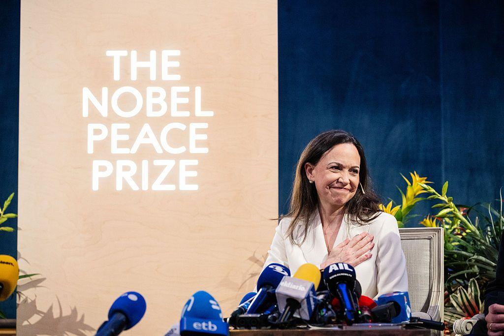 María Corina Machado detrás de unos micrófonos y con un cartel del premio Nobel de la Paz detrás. 