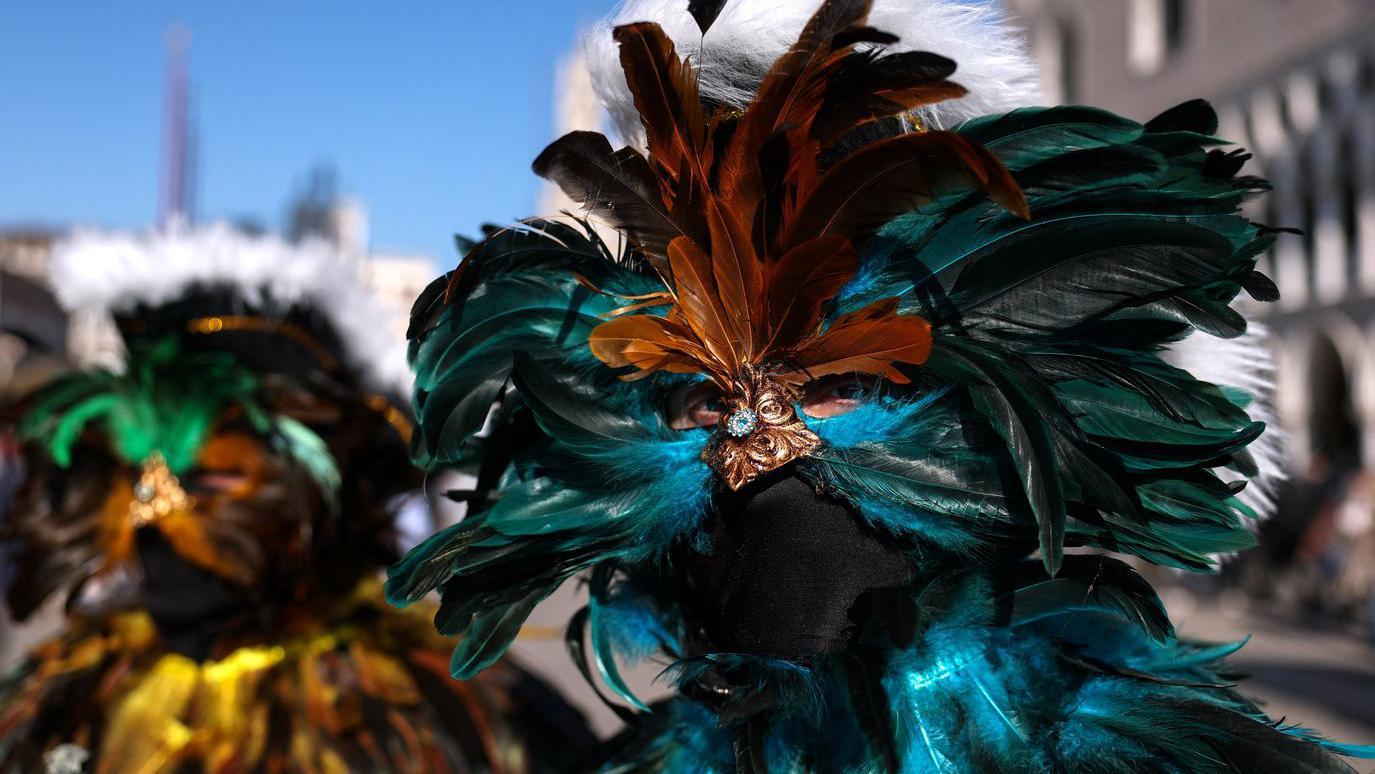 Muškarac drži masku napravljenu od plavo-zelenih pera, vide mu se samo oči