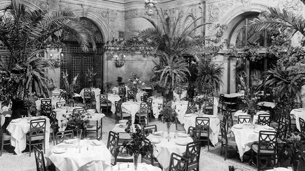 Una imagen en blanco y negro del comedor con mesas redondas vestidas con manteles blancos y un florero en el centro de cada mesa, y palmeras que decoran la sala.