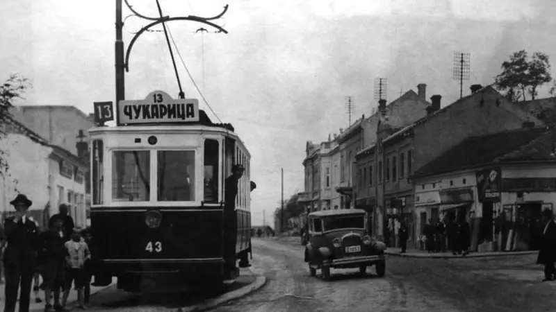 Tramvaj broj 13 na beogradskim ulicama, na crno beloj fotografiji, pored je jedan automobil, sa desne strane a sa leve ljudi