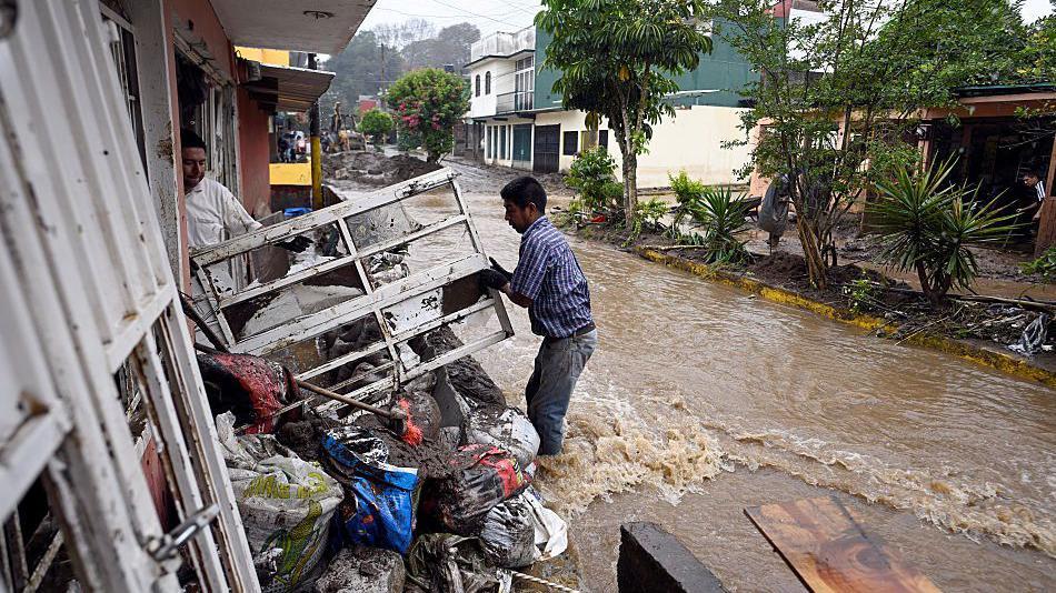 Dos hombres intentan despejar escombros de una casa, mientras uno está parado con el agua hasta las rodillas en una calle inundada