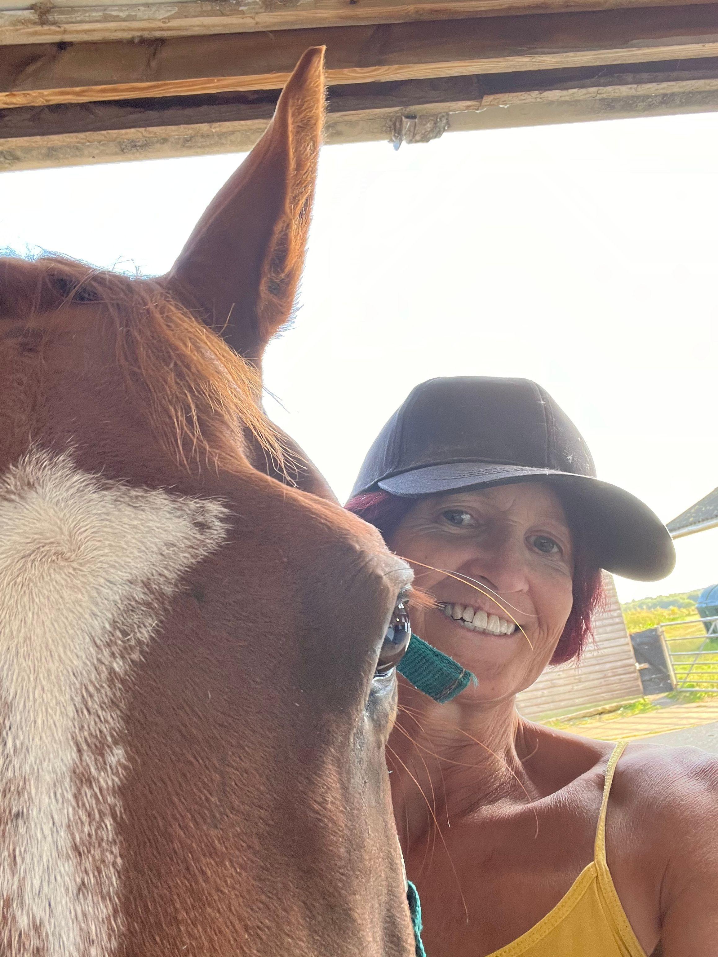 Sue - a woman smiling while wearing a baseball cap - stands next to a horse. The horse is closer to the camera, with only it's eye and forehead visible beside her.
