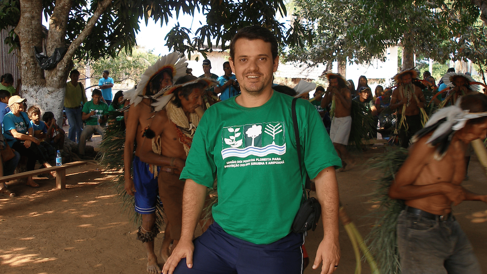 Fonseca sorrindo em área aberta e arborizada; atrás dele, indígenas fazem movimento circular