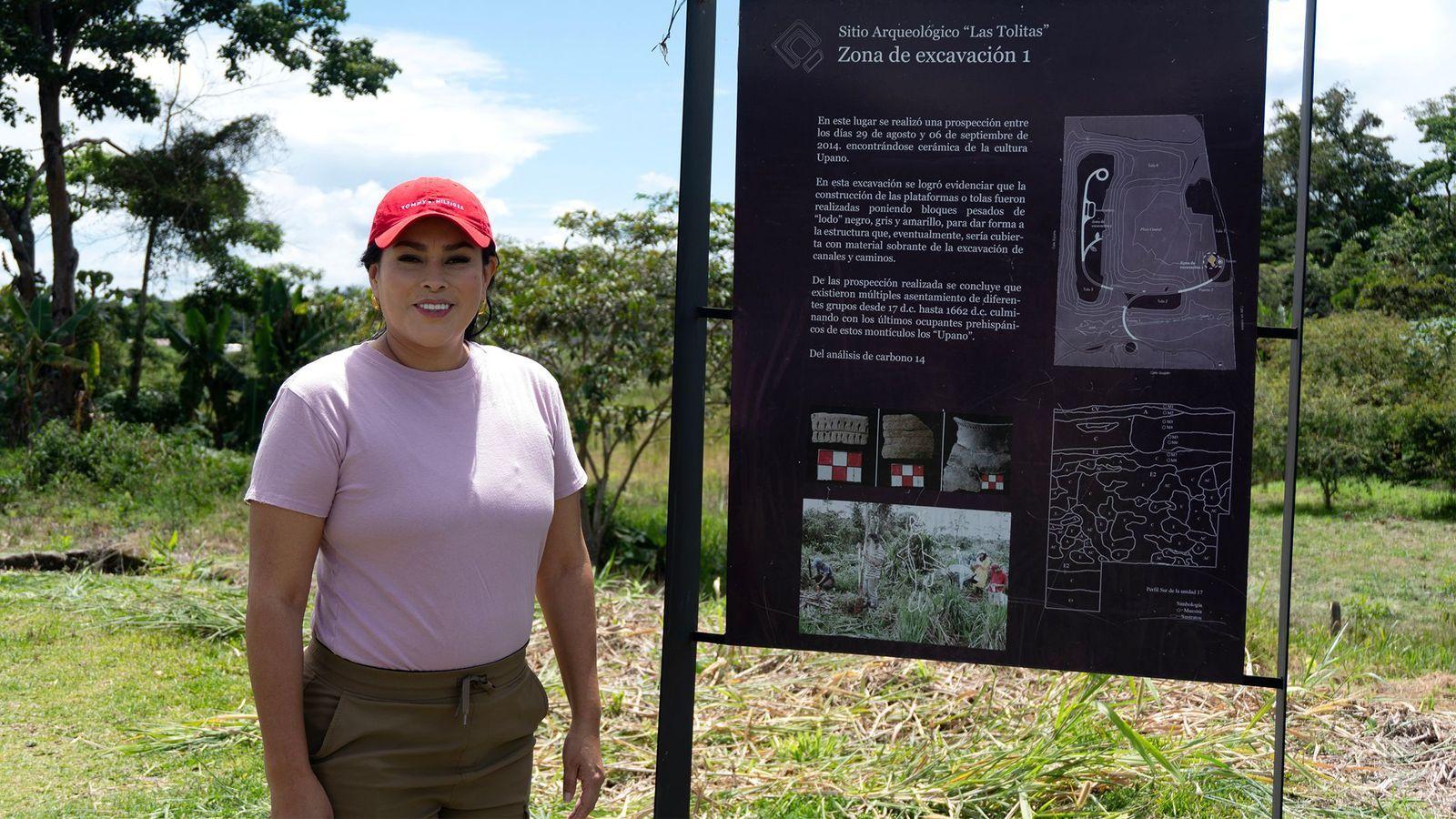 Mujer junto a un cartel informativo de un sitio arqueológico en un entorno rural.