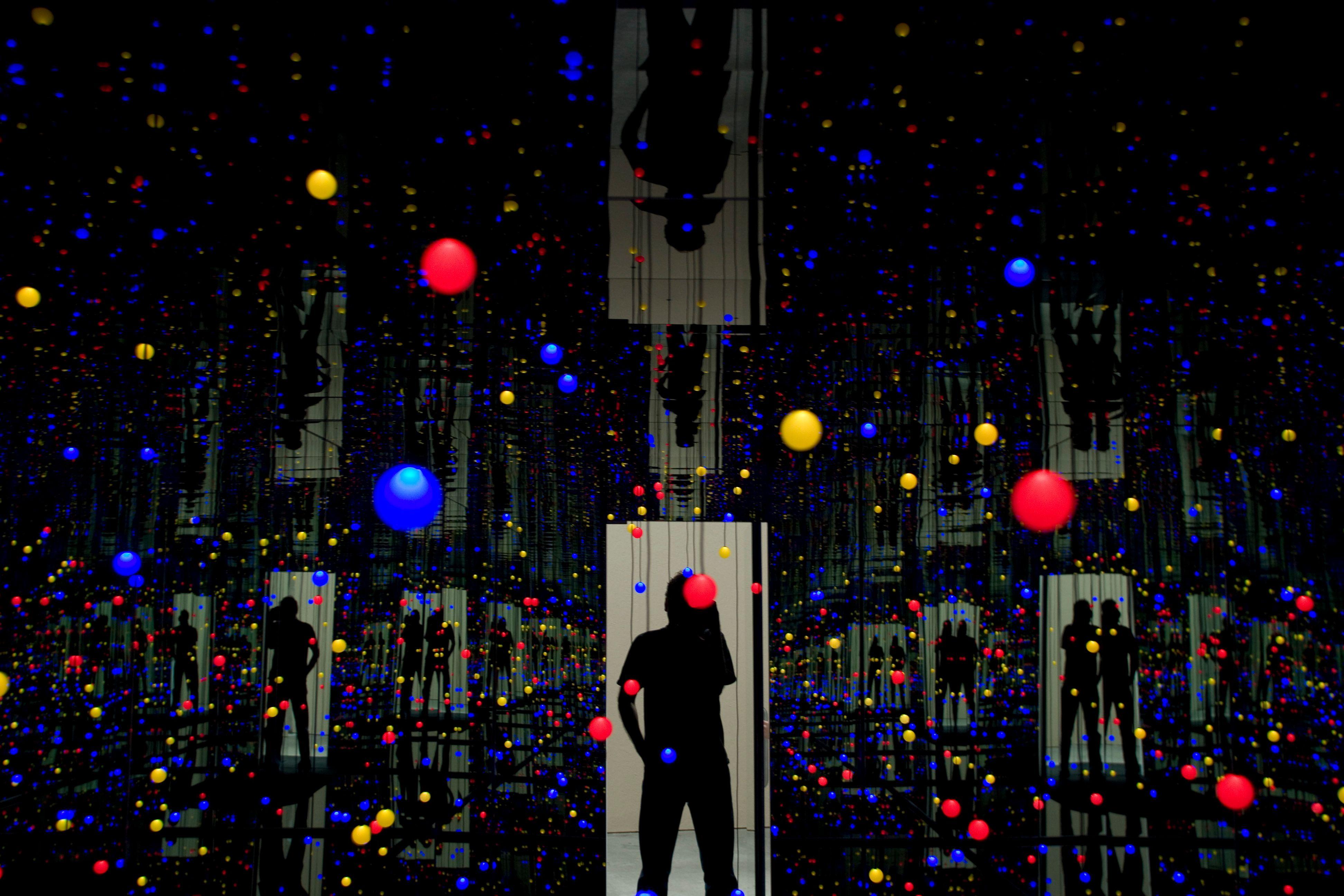 Una instalación de Kusama: un espacio oscuro con muchos pequeños puntos de colores en el aire y reflejos de puertas iluminadas con figuras en silueta.