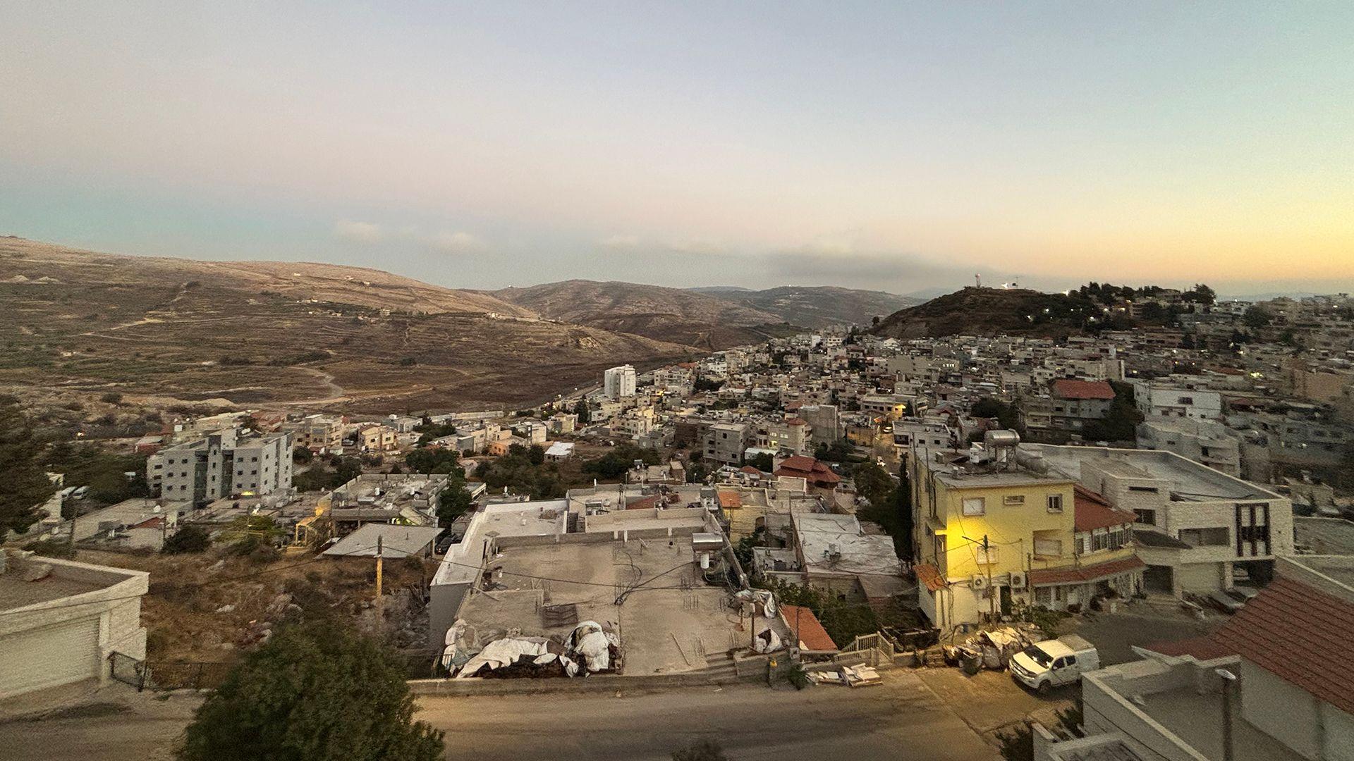 صورة من أعلى لبلدة صغيرة يظهر فيها الكثير من المباني الخرسانية المنخفضة. هناك تلال في الخلفية. السماء ملبدة بالغيوم لونها رمادي-أزرق، ويلوح في الأفق أثر خافت لأشعة الشمس.