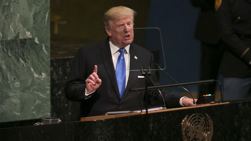 Donald Trump habla ante la Asamblea General de la ONU. Viste un traje oscura, camisa blanca y corbata celeste. Apoya la mano izquierda sobre el podio y tiene la derecha levantada con el dedo índice señalando hacia arriba.