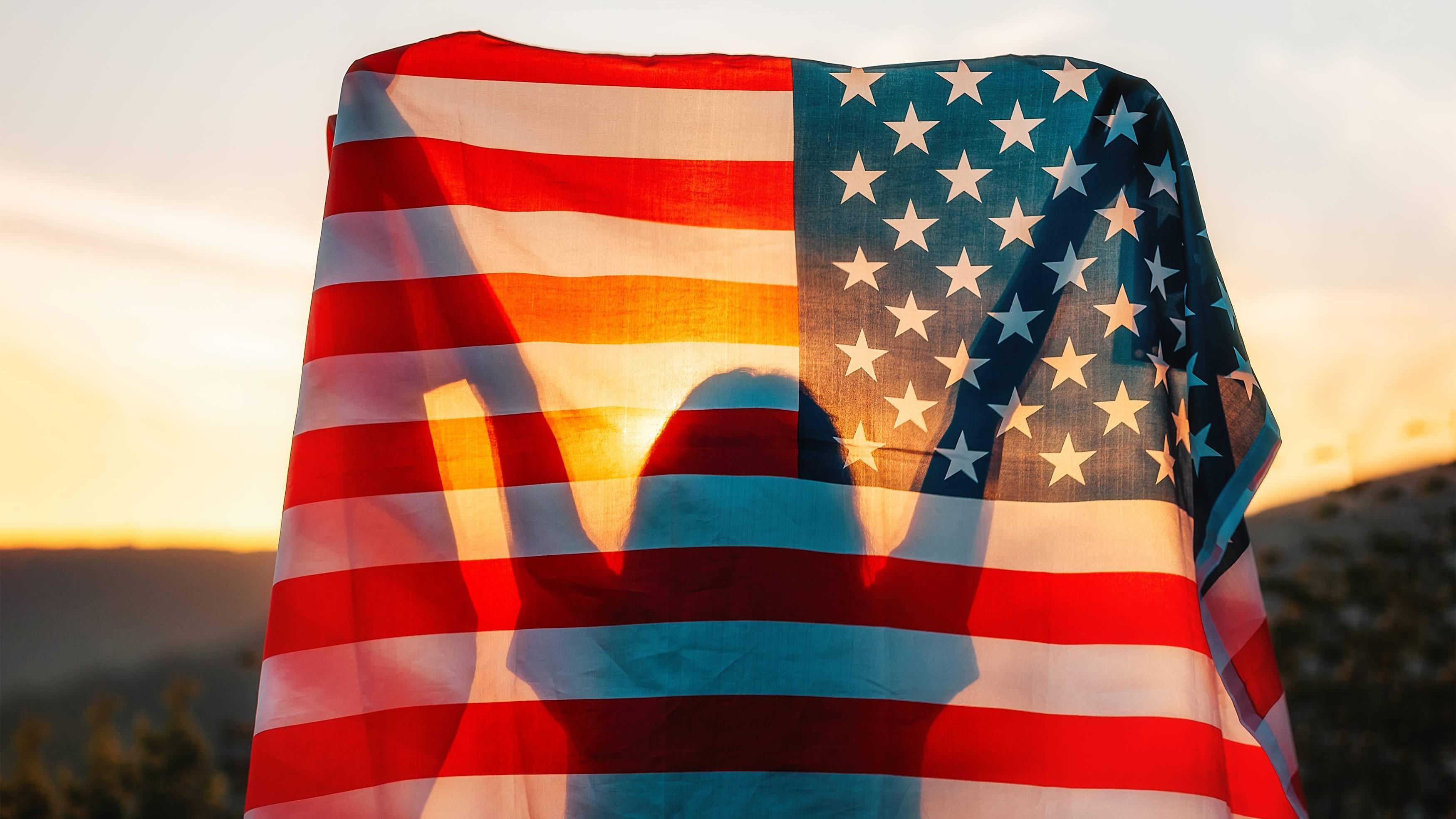 Silueta de una persona con los brazos levantados detrás de una bandera de Estados Unidos iluminada por la luz del atardecer.