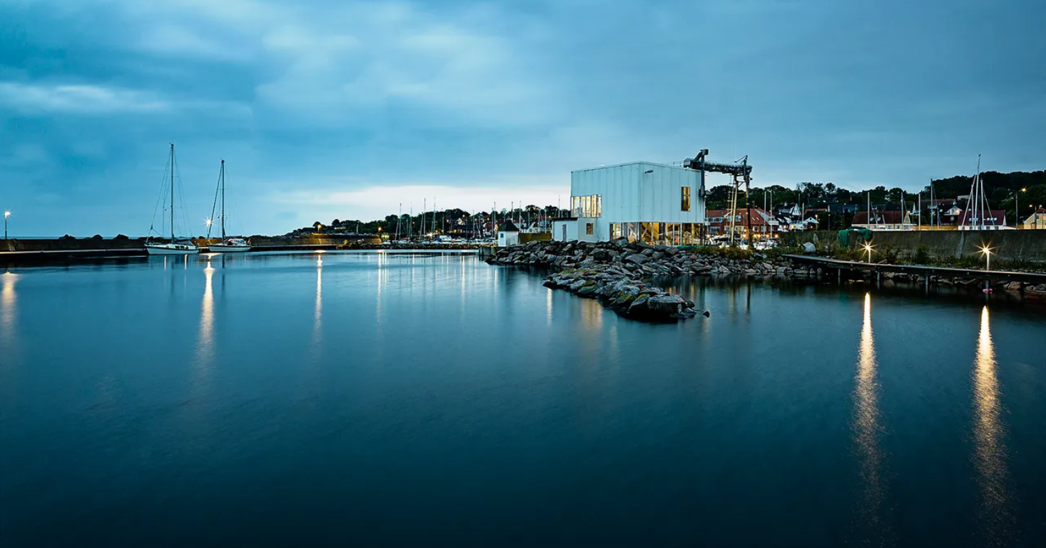 Vista del puerto de Tejn al atardecer, con aguas tranquilas que reflejan las luces, embarcaciones amarradas y un edificio moderno junto a un rompeolas de rocas.
