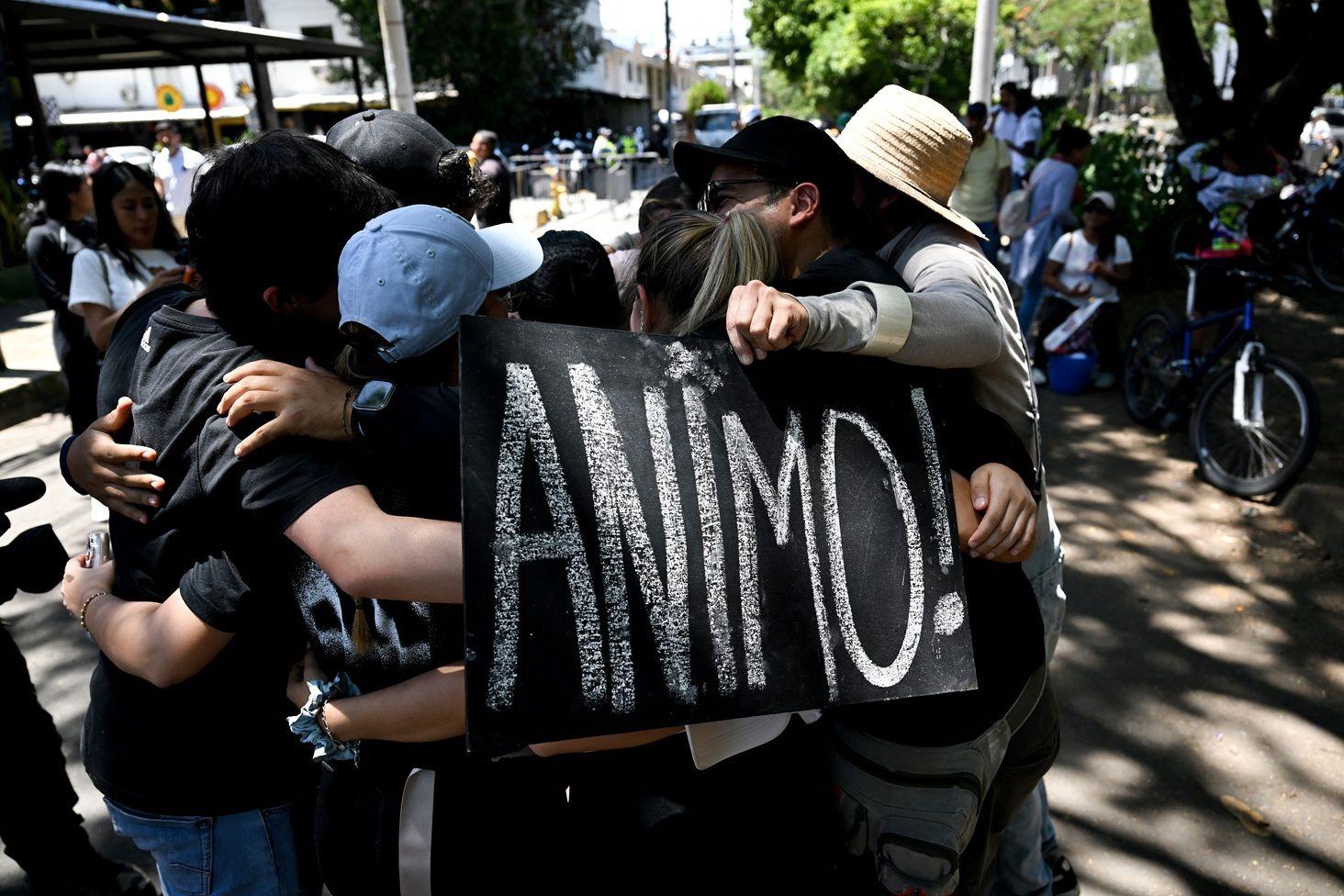 Un grupo de personas se abraza en lo que parece ser una plaza. Una de ellas tiene un cartel negro con letras en blanco que dice: ANIMO.
