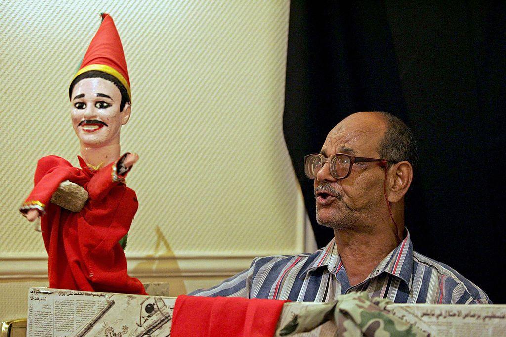 رجل يمسك بأراجوز يرتدي جلباباً أحمر وطرطوراً. 