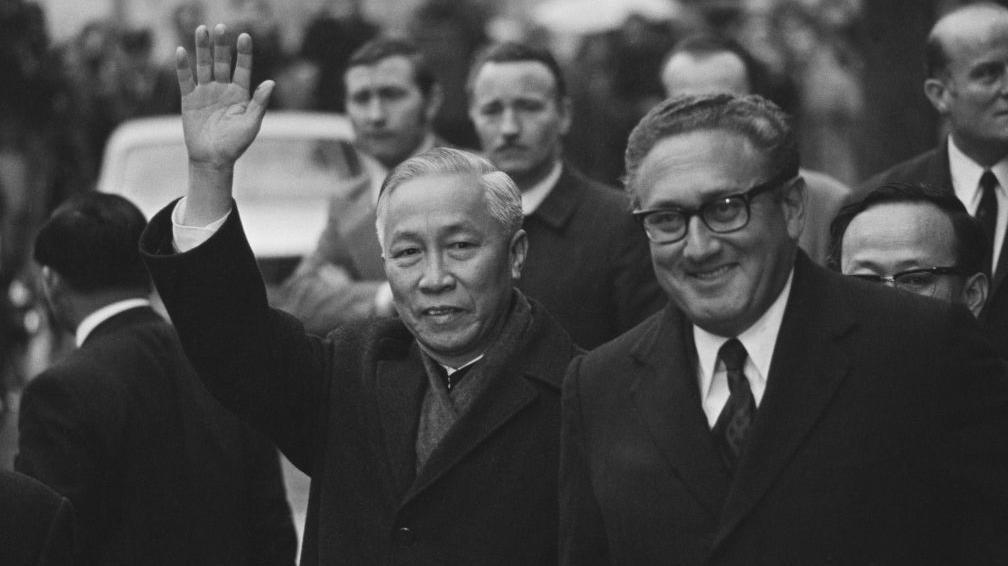 Le Duc Tho levanta una mano al camarógrafo mientras camina junto a Henry Kissinger durante las negociaciones de París.