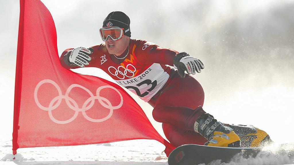 Ryan Wedding en una snowboard con el uniforme rojo de los Juegos Olímpicos de Salt Lake City, gafas de nieve, botas amarillas y un gorro negro. A su lado ondea una bandera roja con el logotipo olímpico en blanco.
