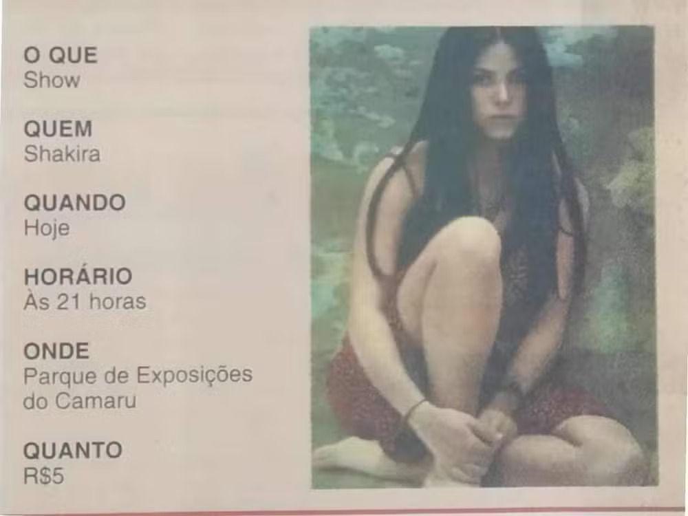 Um cartaz divulga um show de Shakira, com informa&ccedil;&otilde;es organizadas &agrave; esquerda &mdash; &ldquo;O que: Show&rdquo;, &ldquo;Quem: Shakira&rdquo;, &ldquo;Quando: Hoje&rdquo;, &ldquo;Hor&aacute;rio: &agrave;s 21 horas&rdquo;, &ldquo;Onde: Parque de Exposi&ccedil;&otilde;es do Camaru&rdquo; e &ldquo;Quanto: R$5&rdquo;. &Agrave; direita, h&aacute; uma foto da cantora mais jovem, de cabelos pretos, sentada com express&atilde;o s&eacute;ria, em um cen&aacute;rio de tons esverdeados.