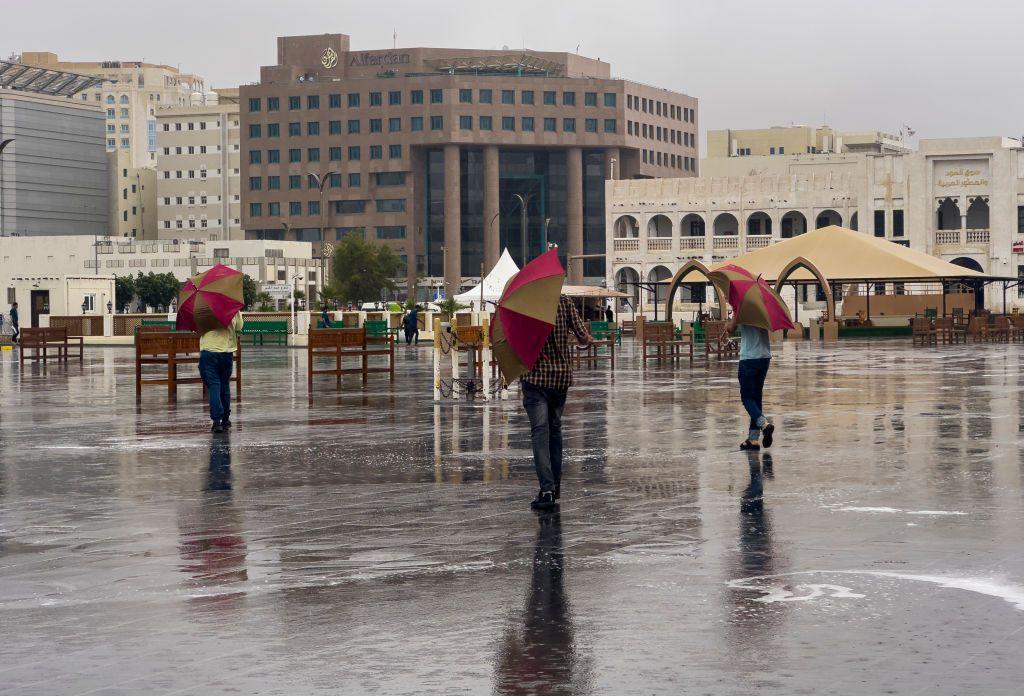 يتجول 3 أشخاص، بمظلات في سوق واقف، خلال يوم ماطر في الدوحة- قطر. تظهر مجموعة من المباني، والأمطار تشكل تجمعاً من المياه.