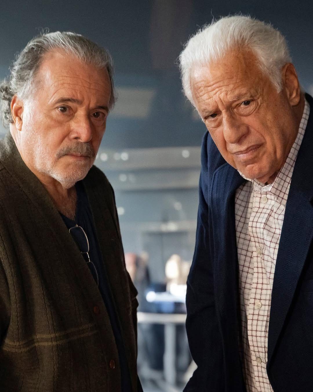 Dois homens idosos encaram a c&acirc;mera com express&atilde;o s&eacute;ria. O da esquerda, Tony Ramos, usa uma camiseta escura com um casaco marrom por cima, e &oacute;culos pendurados na gola; o da direita, Antonio Fagundes, veste camisa xadrez clara sob um blazer azul.