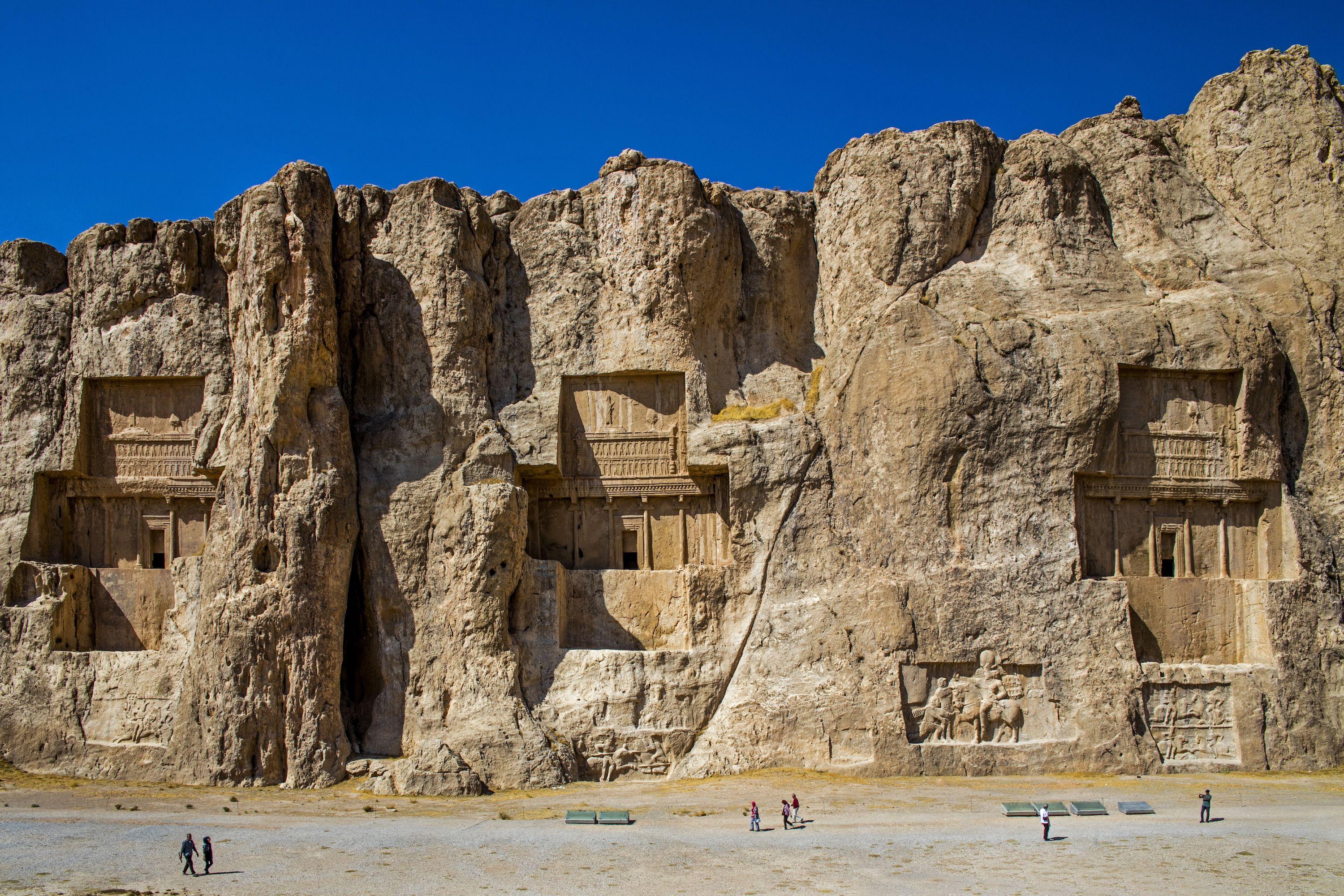 Una antigua necrópolis persa está excavada directamente en una enorme pared rocosa. La piedra caliza presenta portales de formas intrincadas. Los turistas pasan caminando bajo un cielo azul intenso.