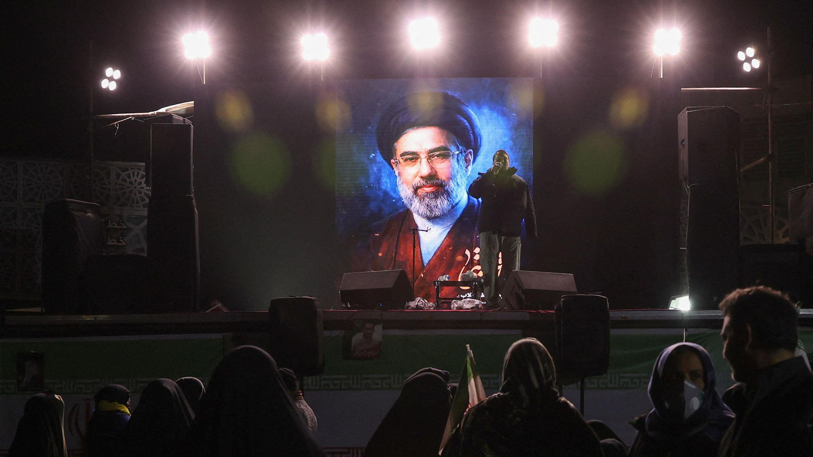 تُظهر الصورة مجتبى خامنئي، المرشد الأعلى الإيراني الجديد، على شاشة مضاءة في الظلام في طهران، حيث عُرضت صورته أمام حشد من الناس. ويقف رجل أمام الشاشة حاملاً ميكروفوناً وبجانبه مكبّر صوت، ويبدو أنه يخاطب الحضور.