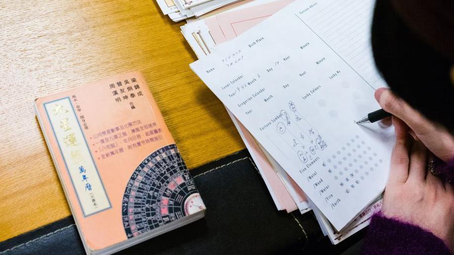 這張照片拍攝於香港一名風水師的辦公室，她正在為美國總統特朗普撰寫豬年運勢的星盤。畫面特寫顯示她的雙手在文件和書本堆上繪製圖表。