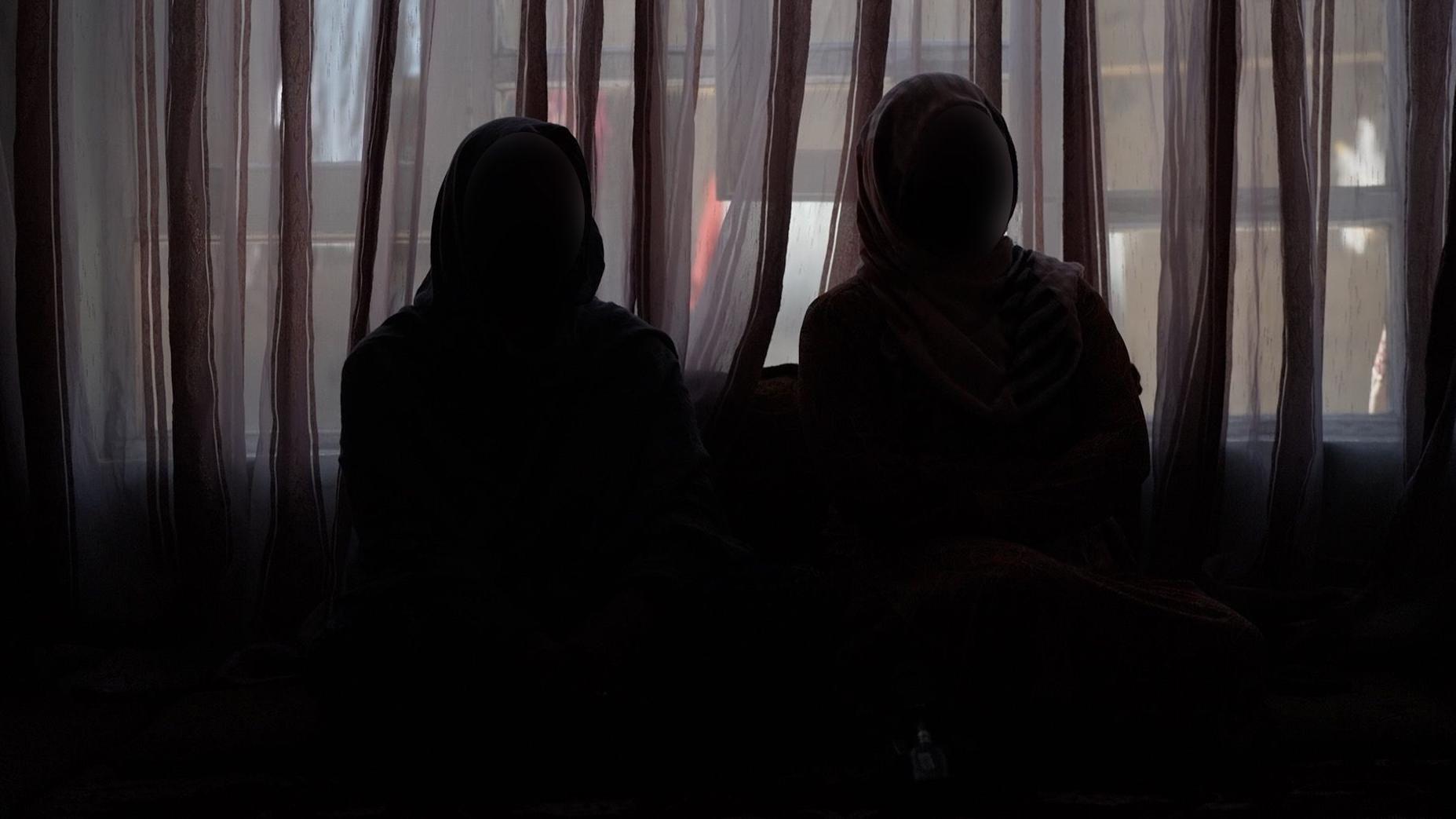 Duas mulheres se sentam no escuro usando hijabs e m&aacute;scaras faciais para proteger sua identidade