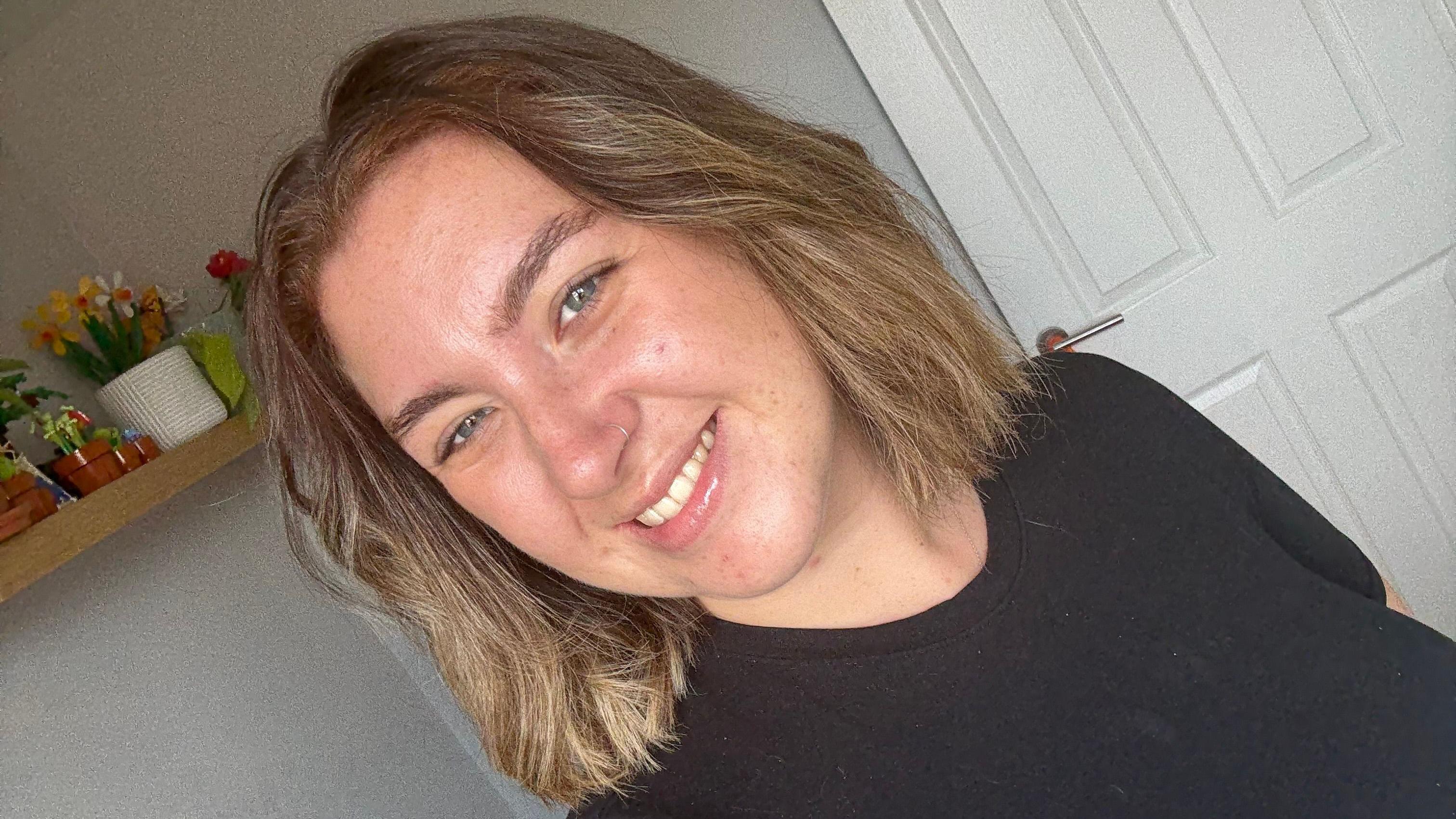 Gabby takes a selfie while smiling. She is wearing a black t-shirt and has shoulder-length brown hair.