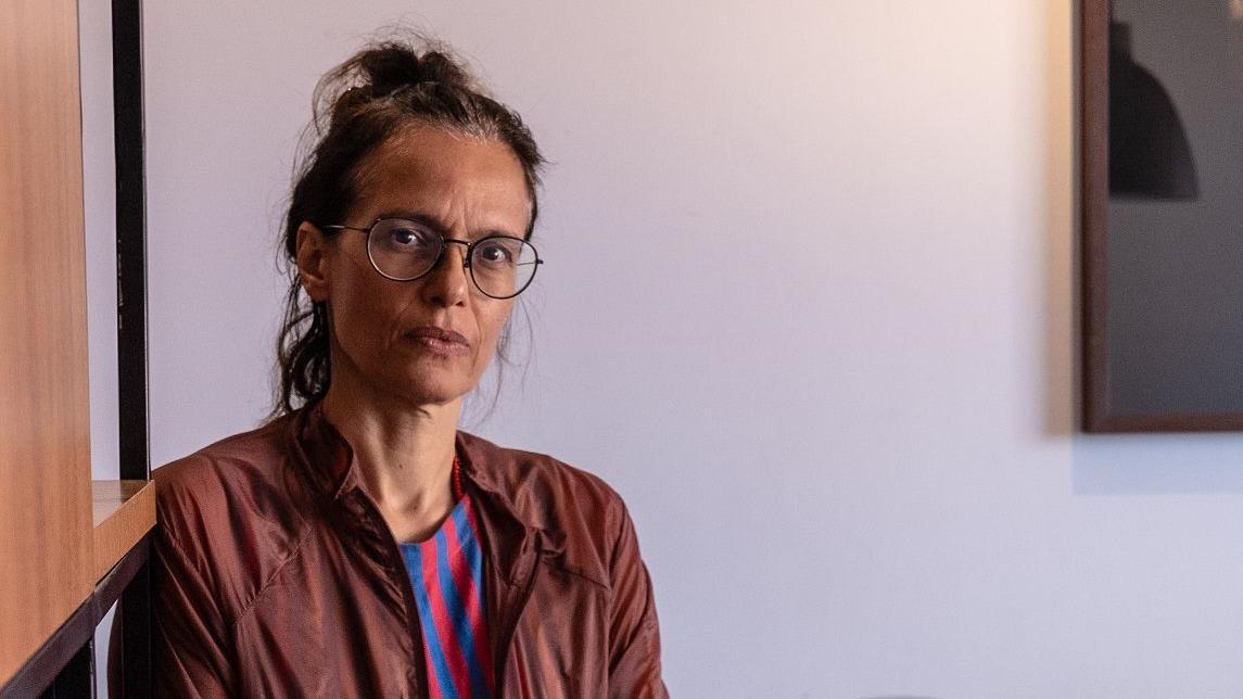 Foto de uma mulher de &oacute;culos de grau, cabelos presos, vestindo uma jaqueta e olhando para a c&acirc;mera. 