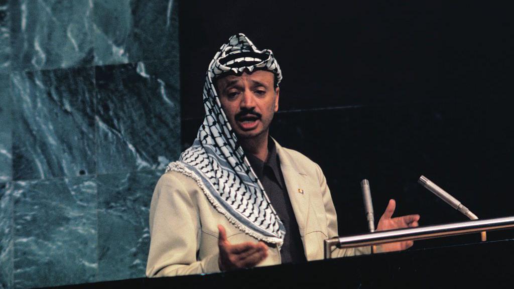 Yasser Arafat habla desde el podio de oradores. Viste un chaqueta beige, una camisa negra y lleva sobre la cabeza una kufiya palestina.