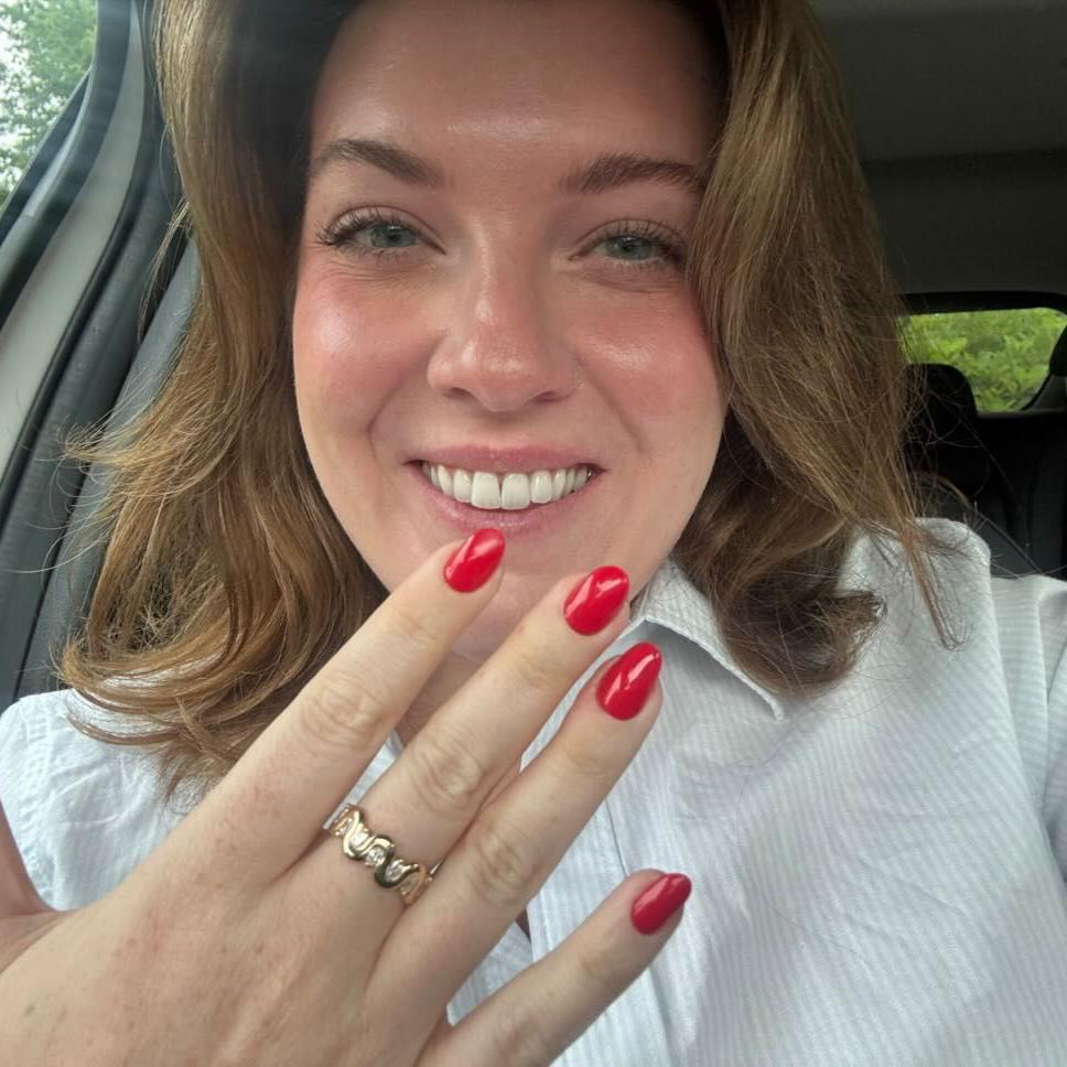 Alex Proie, sentada no carro, mostra sua mão com um anel com design de onda, de ouro e diamante, no seu dedo do meio e sorrindo