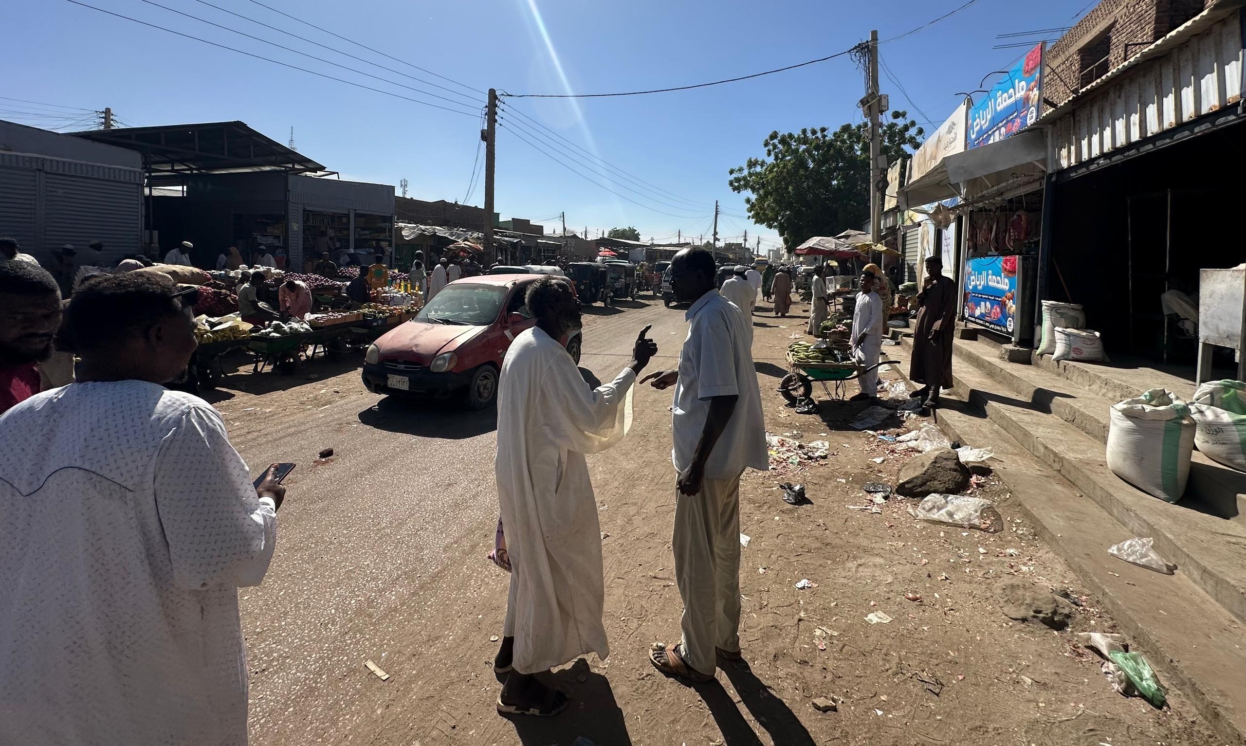 صورة لشارع في مدينة الأبيض به سيارة وباعة جائلون، وسيارة. بينما يقف رجل يرتدي جلباباً أبيض يتحدث إلى رجل يرتدي قميصاً وسروالاً أبيض لونهما رمادي.