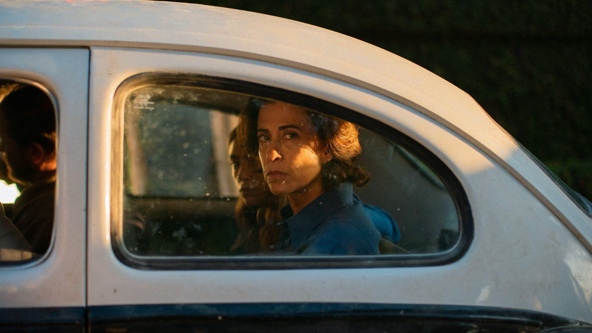 Em cena do filme Ainda Estou Aqui, a personagem Eunice Paiva, interpretada pela atriz Fernanda Torres, &eacute; vista sentada no banco traseiro de um carro antigo, observa pela janela, com express&atilde;o s&eacute;ria.