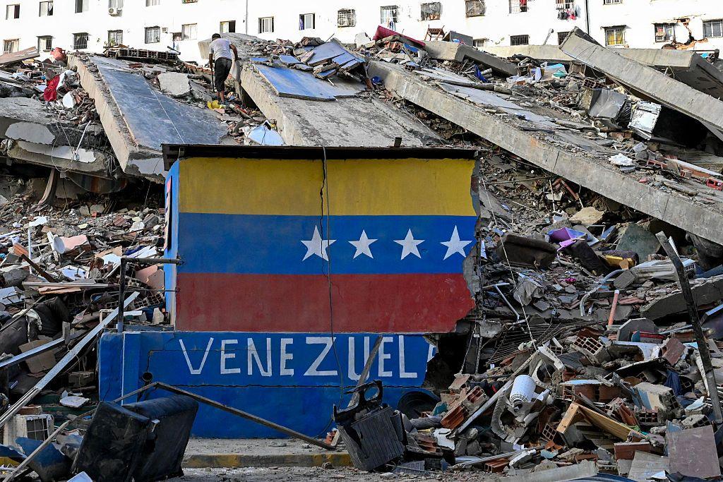 Un muro en el que estaba pintada la bandera de Venezuela yace roto frente a un edificio desplomado por los terremotos en La Guaira, 26 de junio 2026