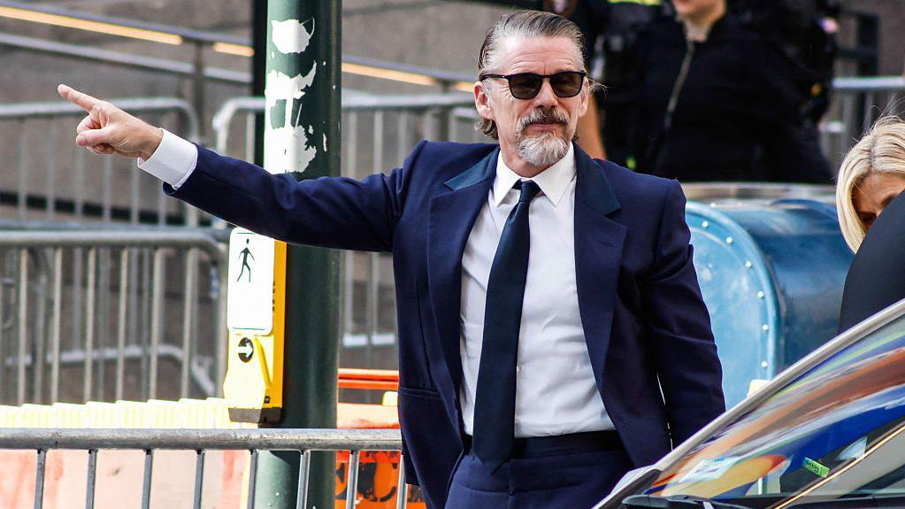 Ethan Hawke aparece vestindo um terno azul-marinho. Ele est&aacute; com o bra&ccedil;o estendido e o dedo apontado