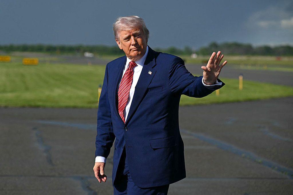 ترامب ملوحاً بيده بعد نزوله من طائرة. الطائرة لا تظهر في الصورة التي التقطت نهاراً. ترامب يرتدي بدلة رسمية كحلية اللون وربطة عنق حمراء مخططة باللون الكحلي وقميص أبيض. يظهر خلفه عشب أخضر.