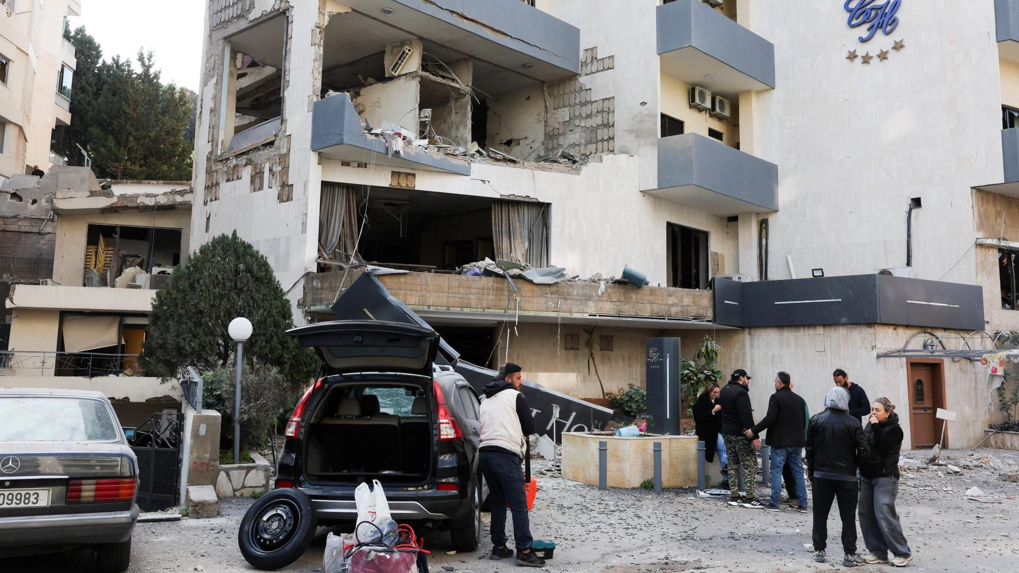 V&aacute;rias pessoas est&atilde;o do lado de fora de um pr&eacute;dio destru&iacute;do, com as janelas estilha&ccedil;adas