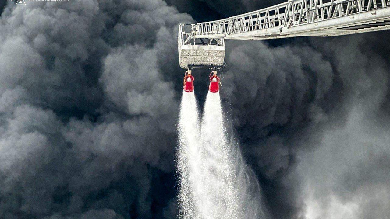 Gust, tamnosivi dim kulja iz naftnog čvorišta Brodi dok vatrogasci gase nevidljivi požar sa dva creva sa produženih merdevina.