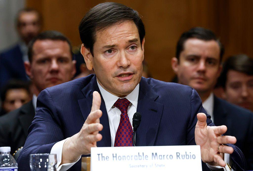 Marco Rubio viste un traje oscuro, camisa blanca y corbata vino tinto. Está sentado mientras habla y hace gestos con las manos.