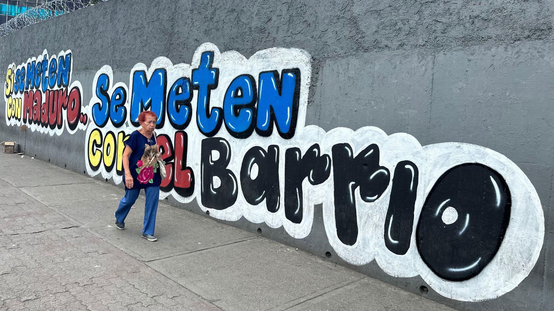 En una pared exterior gris de doble altura con alambre de púas en lo más alto, se extiende un letrero que dice "Si se meten con Maduro, se meten con el barrio", en los colores de la bandera venezolana. Una mujer camina junto al mural.