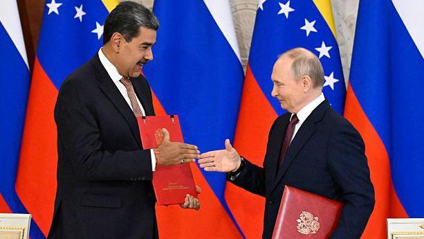 Nicolas Maduro de pie y vestido con traje oscuro extiende su mano derecha hacia Vladimir Putin, quien corresponde al gesto. Ambos sostienen en sus manos izquierdas unas carpetas de cuero rojas. Al fondo se ven las banderas de Rusia y Venezuela.