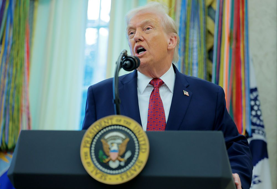 Trump falando ao microfone diante de palanque de presidente dos EUA; ele está dentro de sala